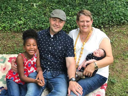 Couple sit on a blanket with their African American daughter and their newly adopted biracial baby thankful for Mardie making adoption her life's work