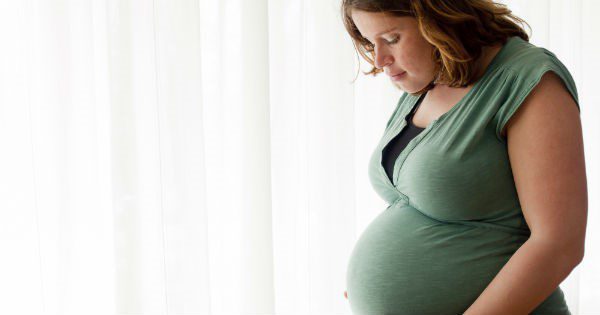 Young pregnant woman in her third trimester standing by a window, looking down at her belly
