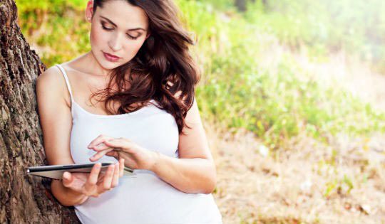 Pregnant woman leaning against a tree and looking at her iPad