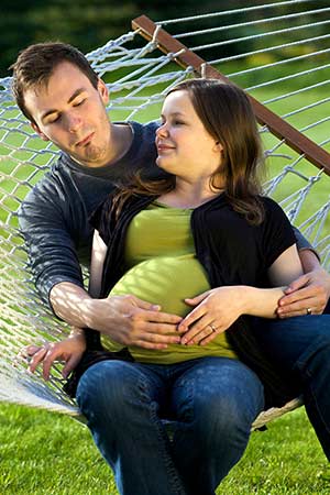 Young couple sit together in hammock, boyfriend touches pregnant girlfriend’s belly to feel a kick