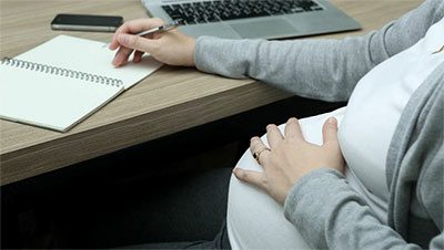 Close up of a pregnant woman writing in a notebook with a laptop nearby