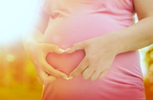 Pregnant woman holding hands in heart shape over stomach with field in the background.