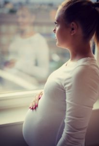 Thoughtful pregnant woman looks out the window home holding hand on tummy thinking about adoption