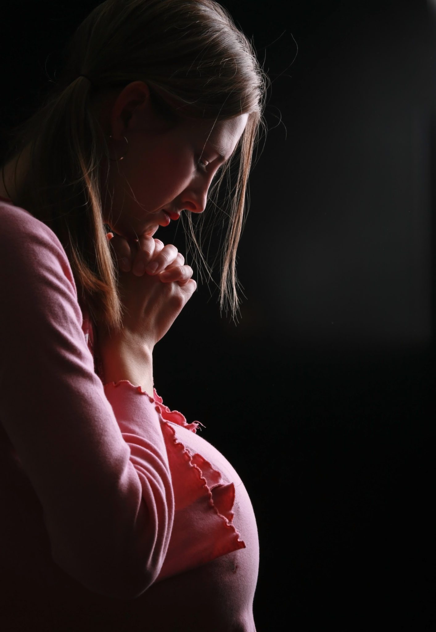 Pregnant woman in a dark room with her hands folded in prayer