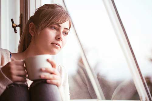 A young woman sits near a window holding a cup of coffee thinking about adoption