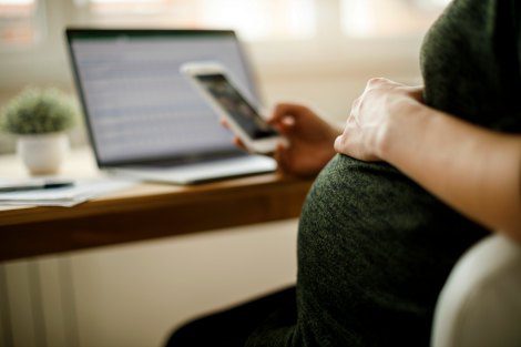 pregnant woman looks at cell phone, laptop in background