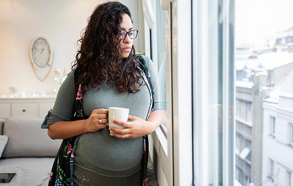 Pregnant woman standing near window with cup of coffee contemplating putting child up for adoption