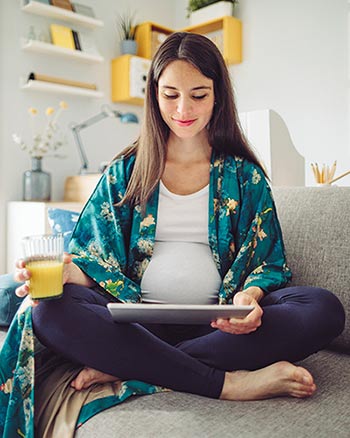Young pregnant woman looking at a tablet while sitting cross-legged on her sofa