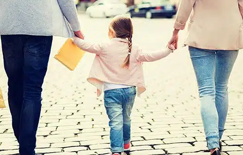 Father and mother hold their daughter's hands as they walk across a parking lot
