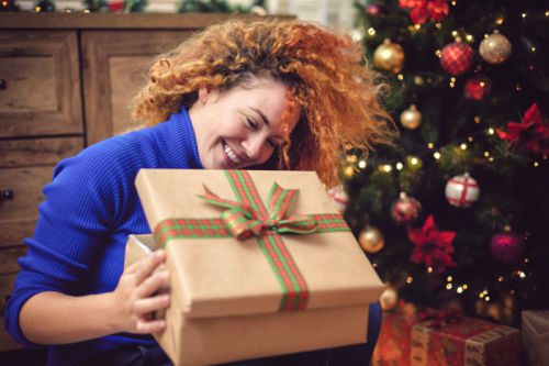 Young woman seated by Christmas tree opening one of the gifts from our birth mother gift ideas list