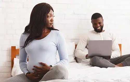 Pregnant woman sitting on her bed, watching her boyfriend and thinking about how to put a baby up for adoption