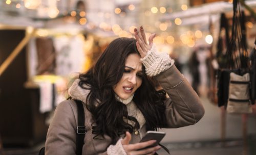 Young woman looking at her phone and holding her head while standing on the street