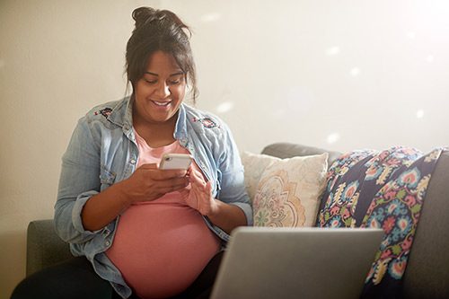 Young pregnant woman using her phone and laptop while sitting in her living room 