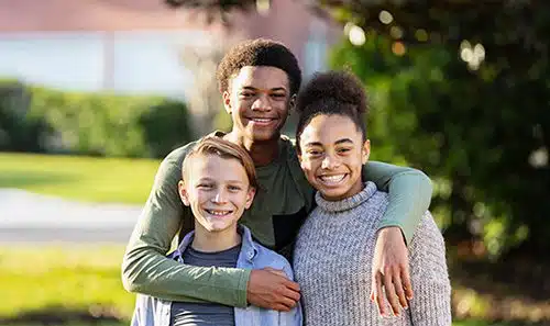 A group of three multi-ethnic children from an adoptive family standing together outdoors in their yard, posing and smiling for the camera