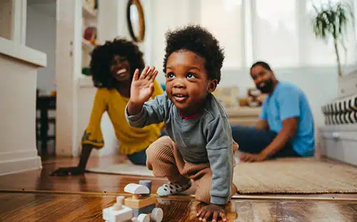 A playful African American toddler playing with a wooden train toy at home while his parents smile at him from the background