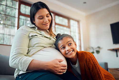 Pregnant mom relaxing with excited, young and curious daughter on sofa