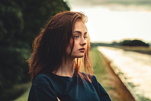 Daydreaming young woman standing alone outdoors close to river in sunset 