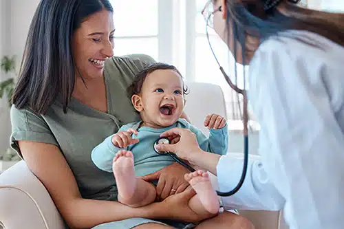 Baby sitting on her mother's lap while being examined by a pediatrician