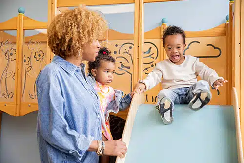 A cheerful toddler boy with Down syndrome explores an indoor play space while his mom and baby sister watch